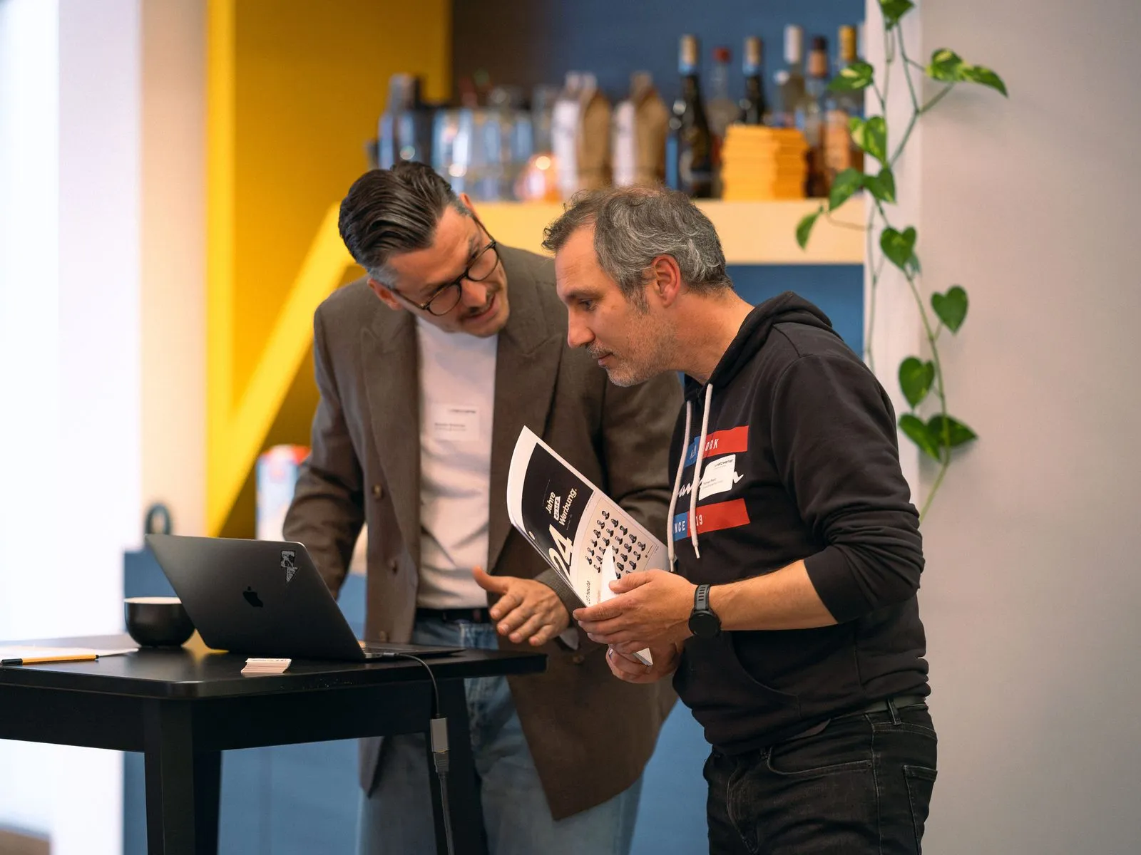 Alexander Schirmer und Thomas Franz beugen sich am Stehtisch über einen Laptop, Thomas Franz hält eine Broschüre