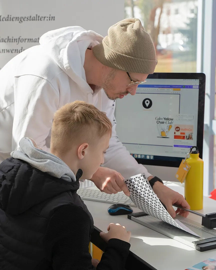 Ein Mitarbeiter der C3 Werbeagentur zeigt einem Jungen, wie man ein selbst gestaltetes Namensschild zuschneidet