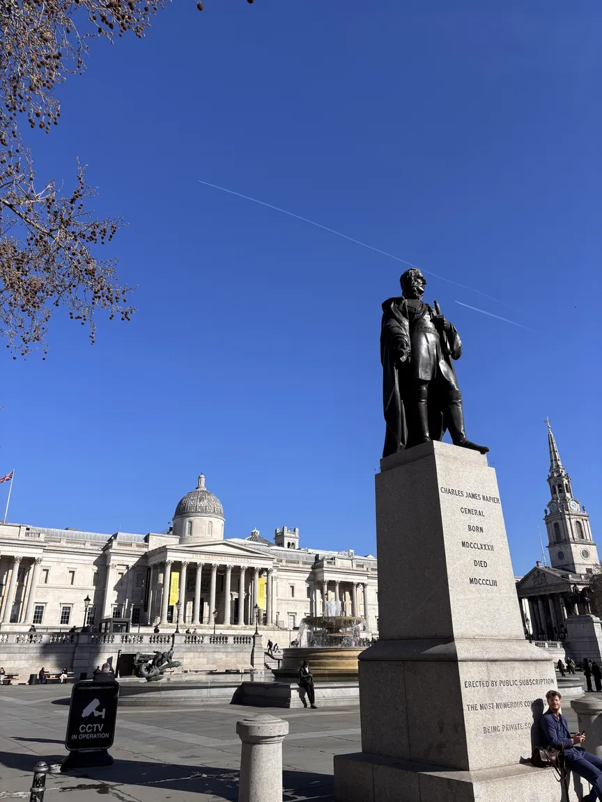 Eine weitwinklige Außenaufnahme des Trafalgar Square in London an einem sonnigen Tag. Im rechten Vordergrund steht die Bronzestatue von Sir Charles James Napier auf einem hohen grauen Steinsockel. Im Hintergrund ist das monumentale Gebäude der National Gallery mit seinen korinthischen Säulen und der zentralen Kuppel zu sehen. Ein strahlend blauer Himmel mit Kondensstreifen eines Flugzeugs spannt sich über die Szenerie.
