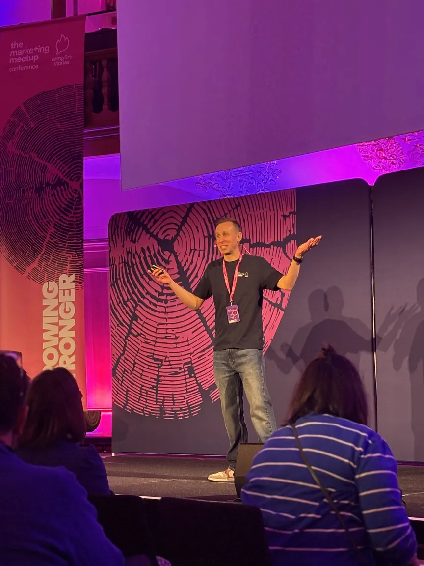 Ein Redner präsentiert auf einer Bühne während der „The Marketing Meetup“-Konferenz. Die Bühne ist in intensives violettes und pinkes Licht getaucht. Im Hintergrund ist eine große Grafik von rosa Baumringen auf dunklem Grund zu sehen, daneben ein Banner mit dem Text „Growing Stronger“. Der Sprecher trägt ein dunkles T-Shirt, Jeans und einen lila Konferenz-Ausweis um den Hals. Im Vordergrund sind die Hinterköpfe der Zuhörer im Publikum erkennbar.