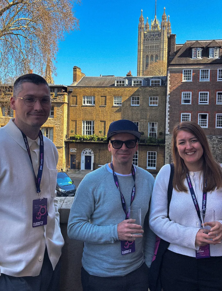 Alexander Schirmer, Christian Reisnecker und Anja Meixner lächeln für ein Porträtfoto auf einem Balkon. Alle drei tragen lilafarbene Lanyards der „Marketing Meetup“-Konferenz. Im Hintergrund erstreckt sich die Londoner Architektur mit historischen Backsteingebäuden und der markanten Spitze des Victoria Tower vom Palace of Westminster unter einem klaren blauen Himmel.