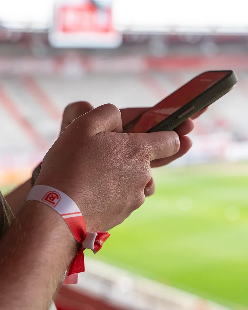 Nahaufnahme von den Händen eines C3-Mitglieds, das vor dem Hintergrund des Fußballstadions ein Smartphone hält. Am Handgelenk ist ein rot-weißes Einlassband mit dem Logo des SSV Jahn Regensburg.