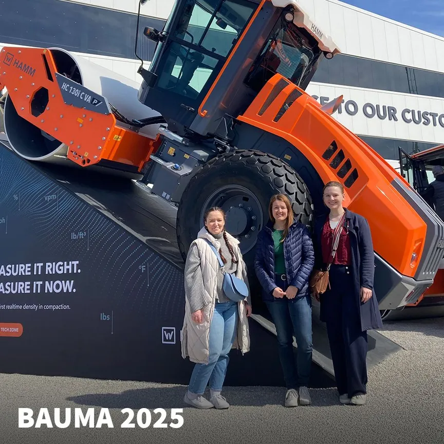 Drei Mitarbeiterinnen der C3 marketing agentur stehen vor einer orangefarbenen HAMM-Baumaschine auf der bauma 2025 in München.