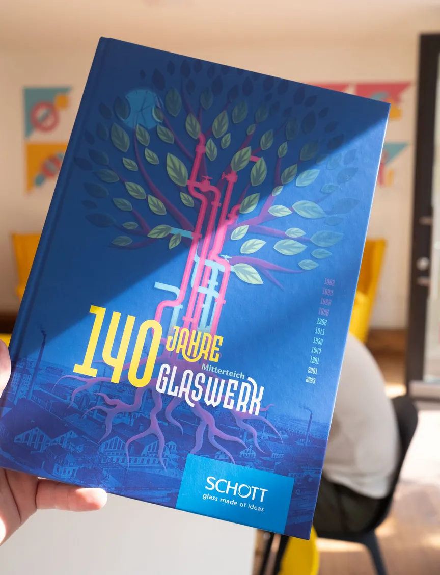 Eine Hand hält ein Buch in die Höhe, auf dessen Cover ein Baum aus Rohren zu sehen ist, dem Titel "140 Jahre Glaswerk Mitterteich" und das Logo von "Schott".