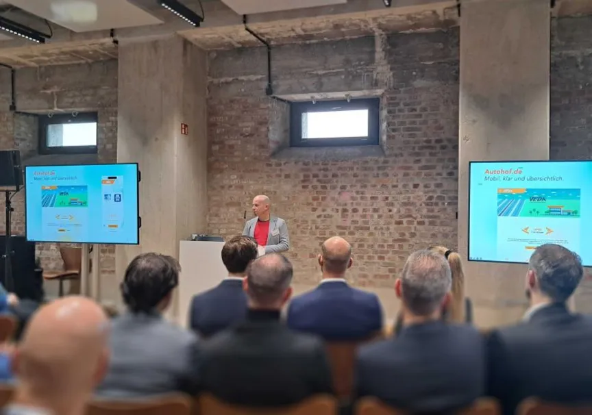 Christian Reisnecker steht an einem Rednerpult im Bergson Kunstkraftwerk München und präsentiert das neue Web- und Markenprojekt für die Vereinigung der deutschen Autohöfe (VEDA). Zwei große Bildschirme flankieren die Bühne, das Publikum verfolgt aufmerksam die Präsentation.